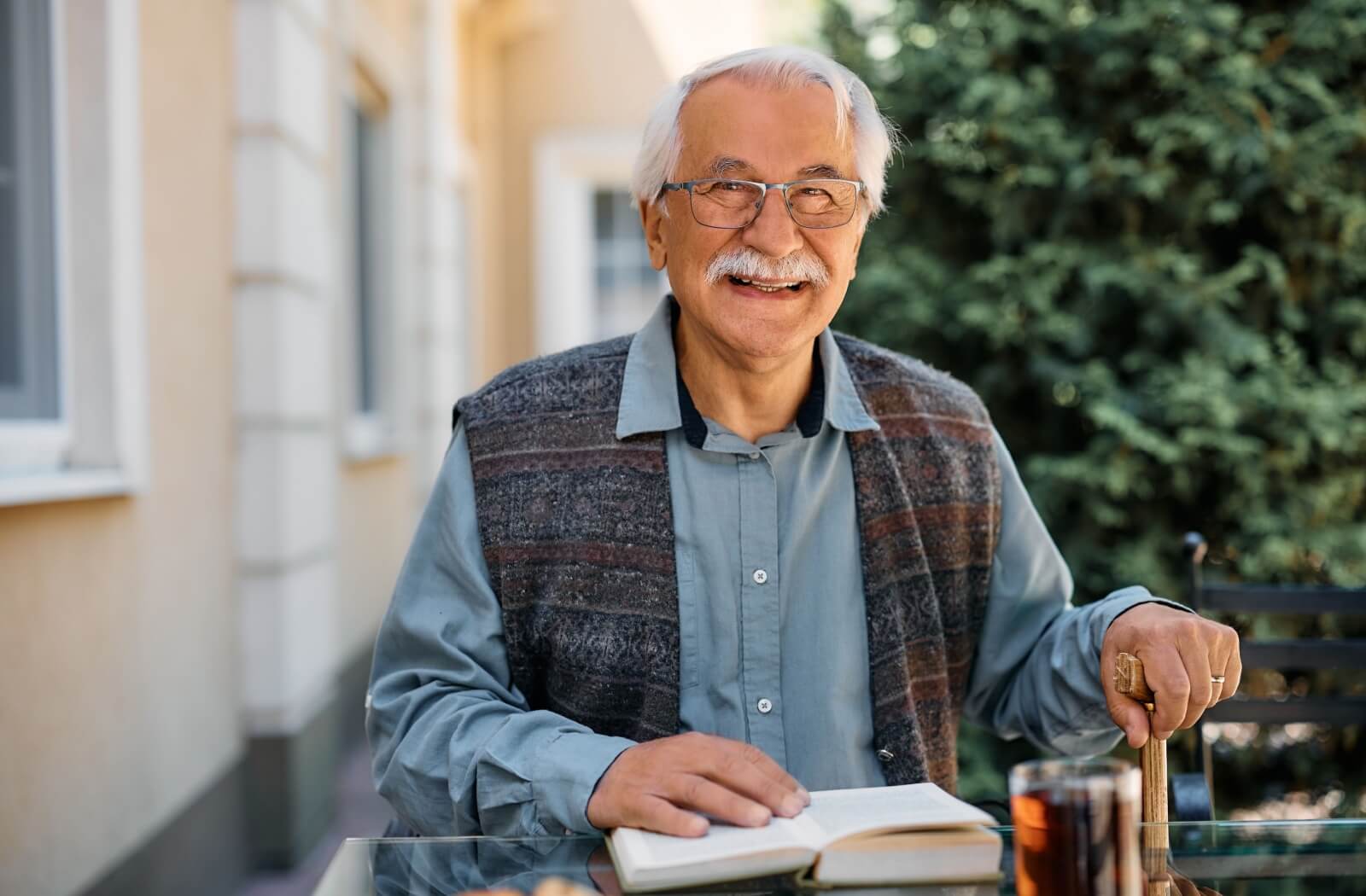 A happy senior reads a book and enjoys their time in an outside common area in their independent living community/