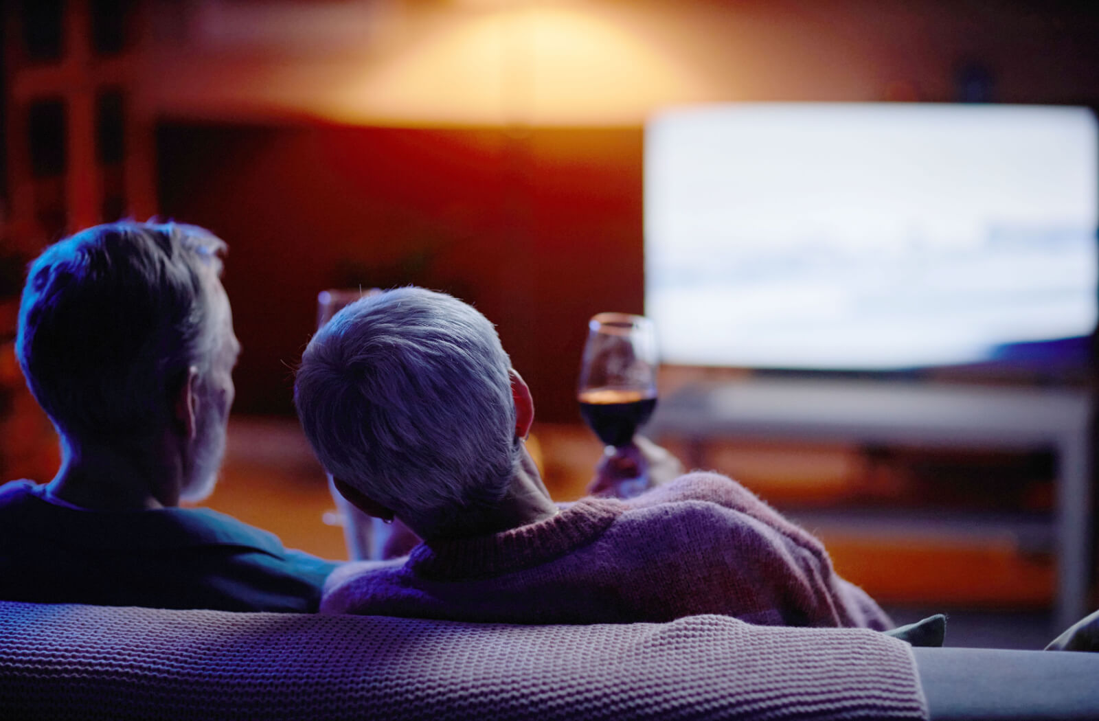 An older couple watching a late-night movie together in a dark living room, raising glasses of wine.