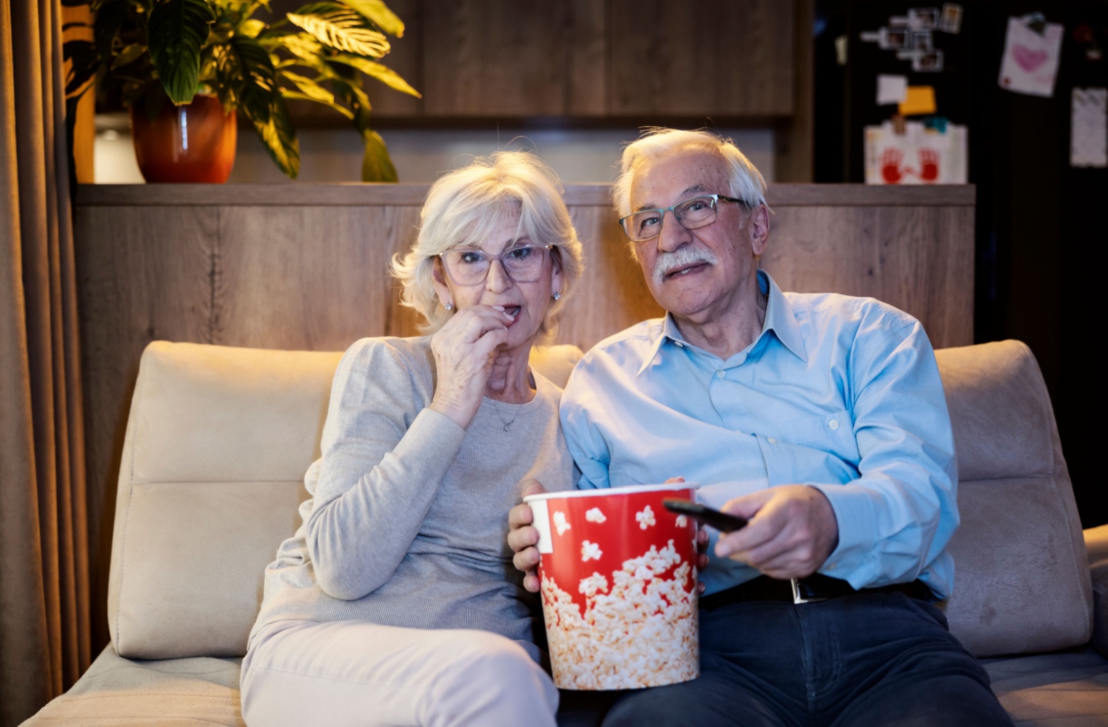 An older couple sitting on the couch with a bucket of popcorn in their living room while watching a movie at night.
