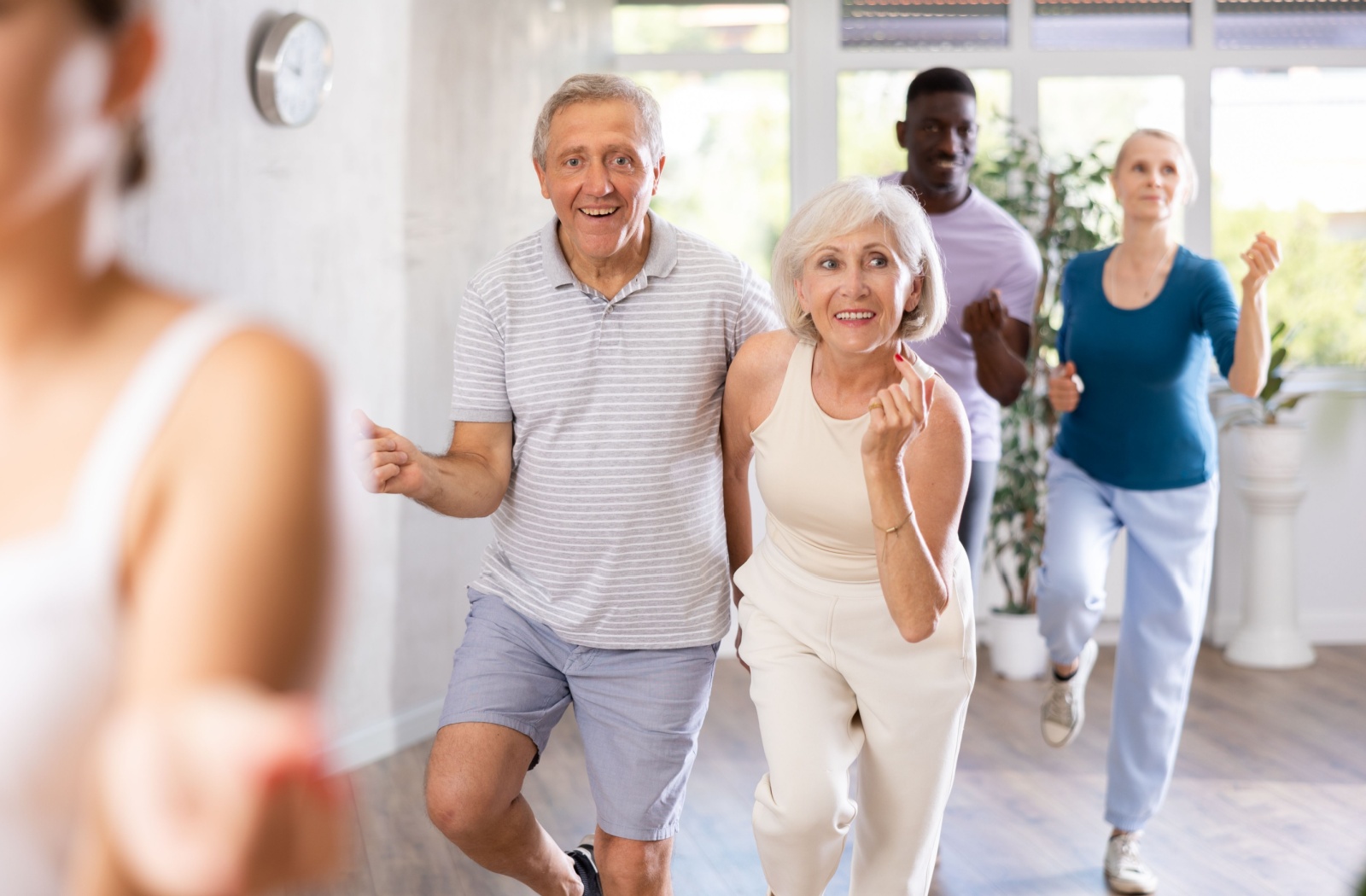 An older couple laughs during a joint line dancing class in a well-lit fitness center
