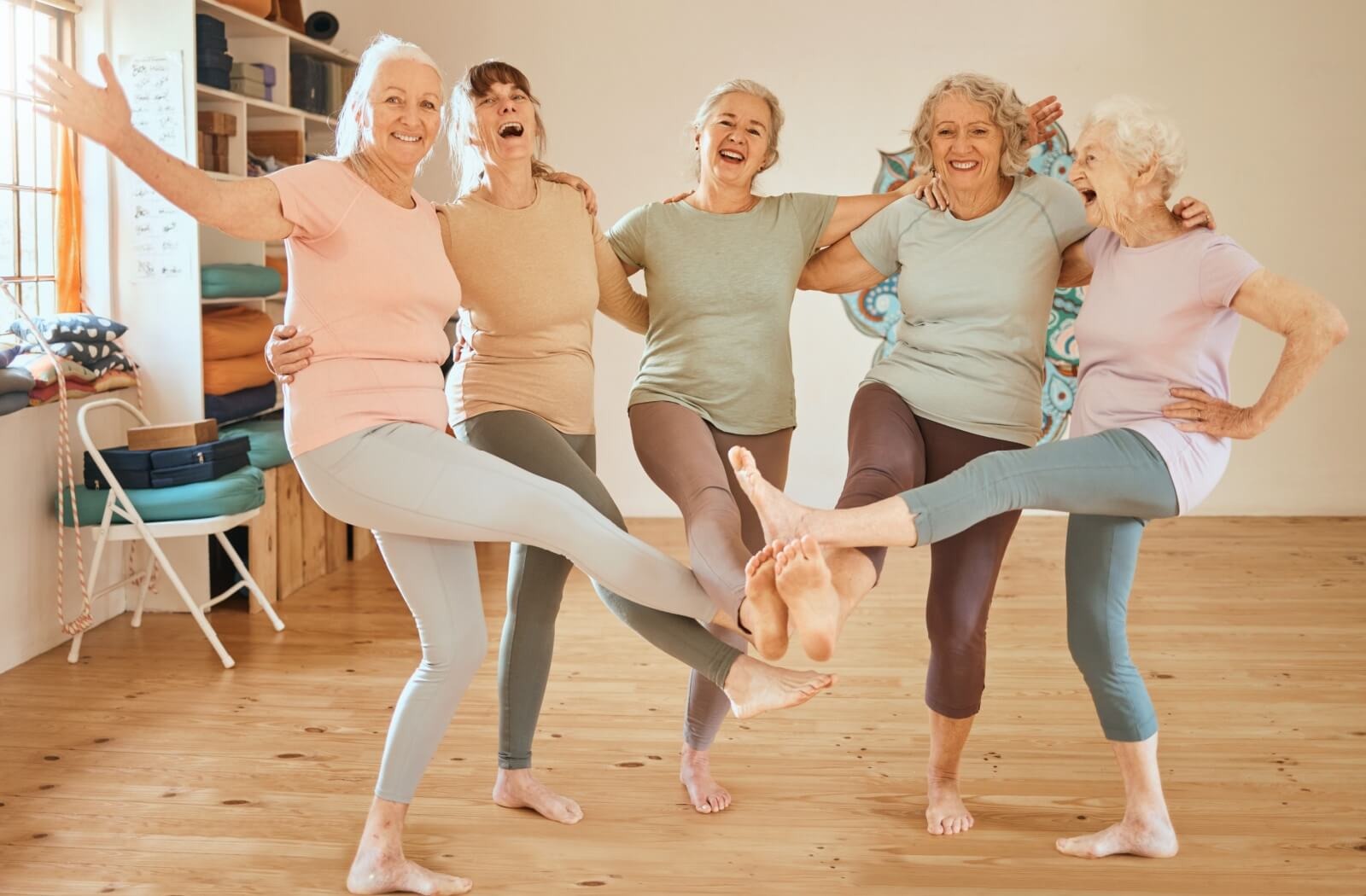 A group of 5 older adults touch their feet together in a circle and laugh while being silly during a line dance practice session