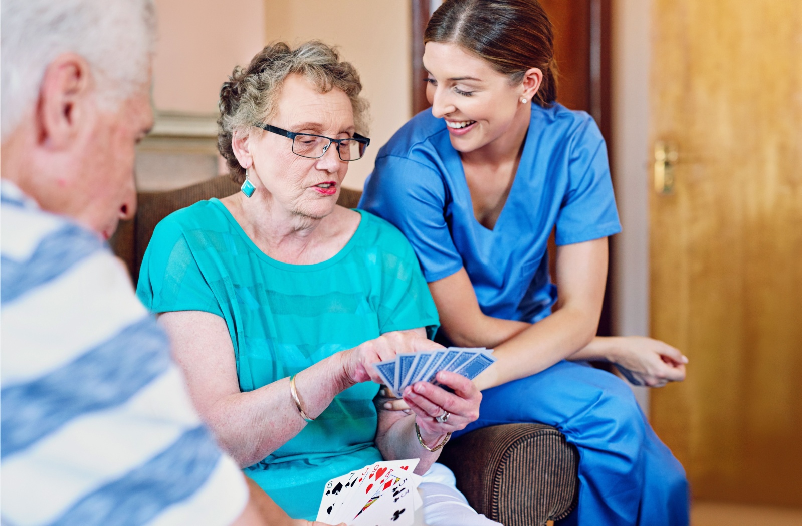 Two senior residents play a game of cards as a caregiver watches.