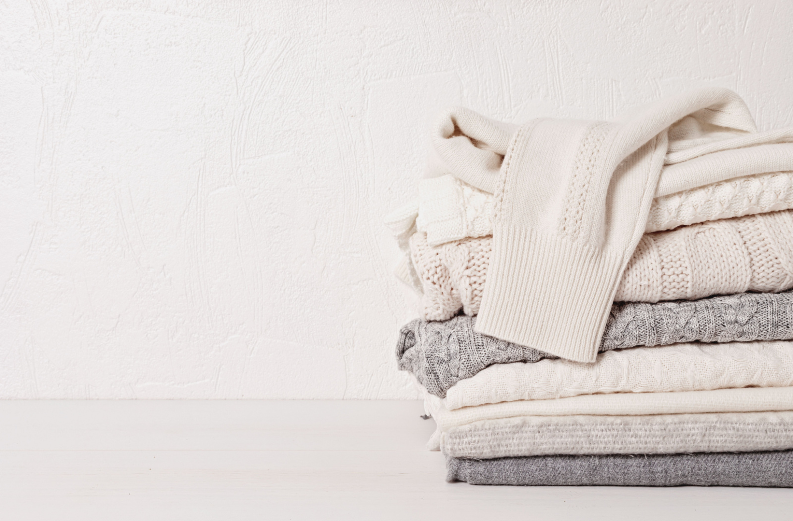 Various garments featuring distinct knitting patterns neatly folded in a stack, showcasing a palette of white and gray hues.