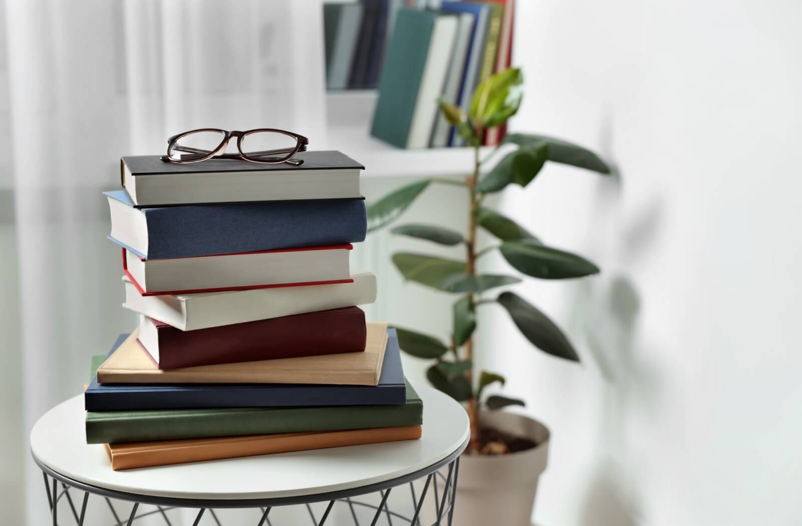 A stack of 10 books sitting on a side table with a pair of black reading glasses resting on top of them and a plant in the background.