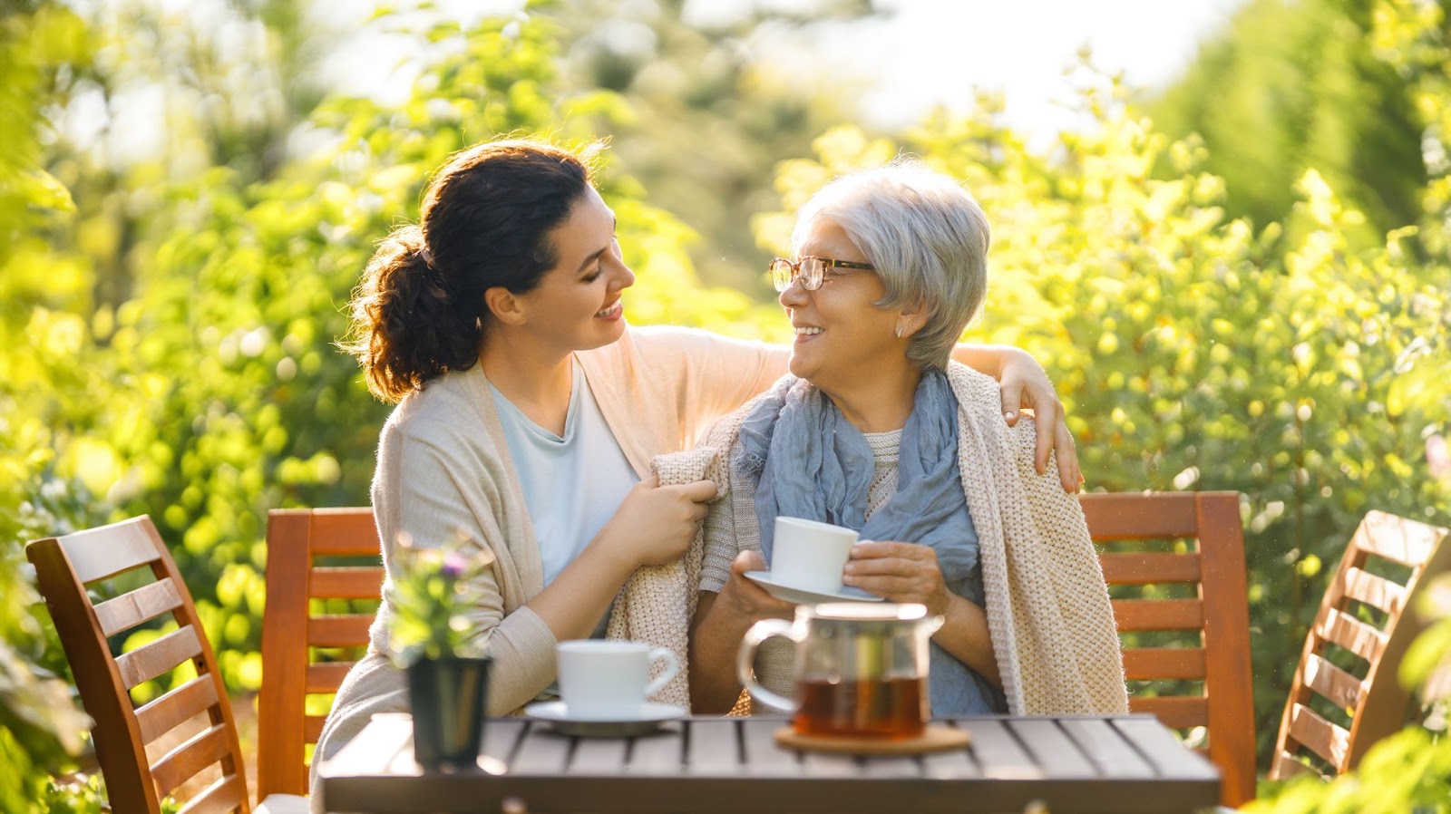 An adult child and her elderly parent smiling and sharing a cup of tea outdoors.