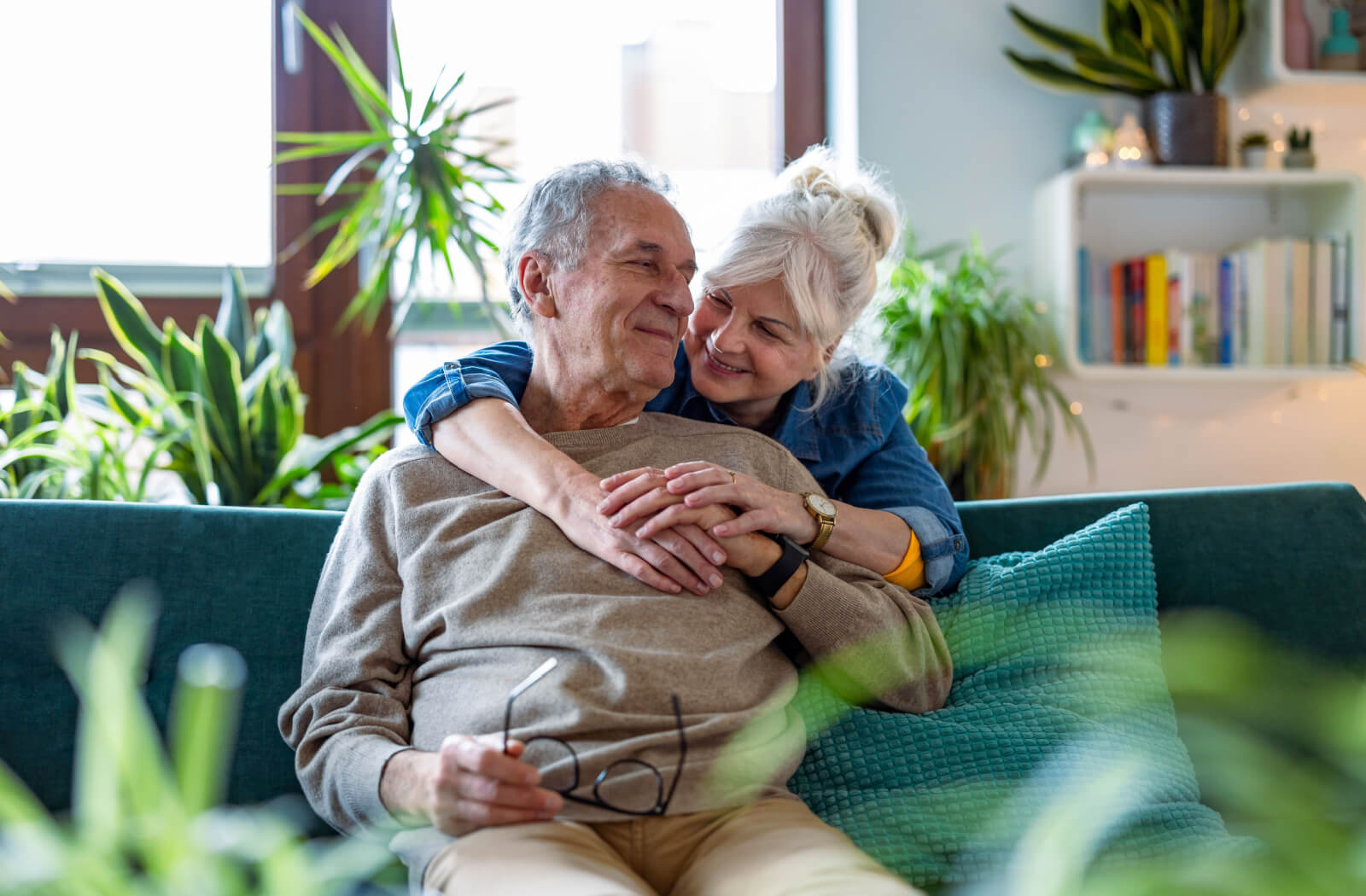 An older adult in senior living hugging their partner from behind in a sunlit room surrounded by plants.