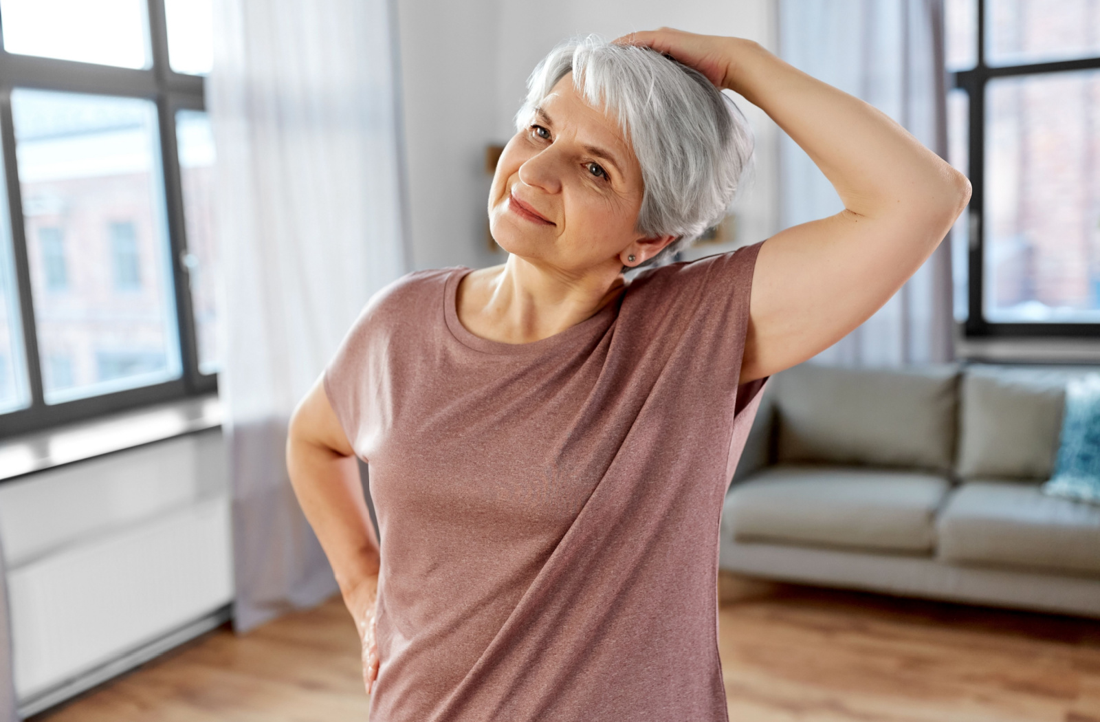 A senior woman gracefully holds her head and gently tilts it to the right, engaging in a neck stretching exercise within the comfort of her home.