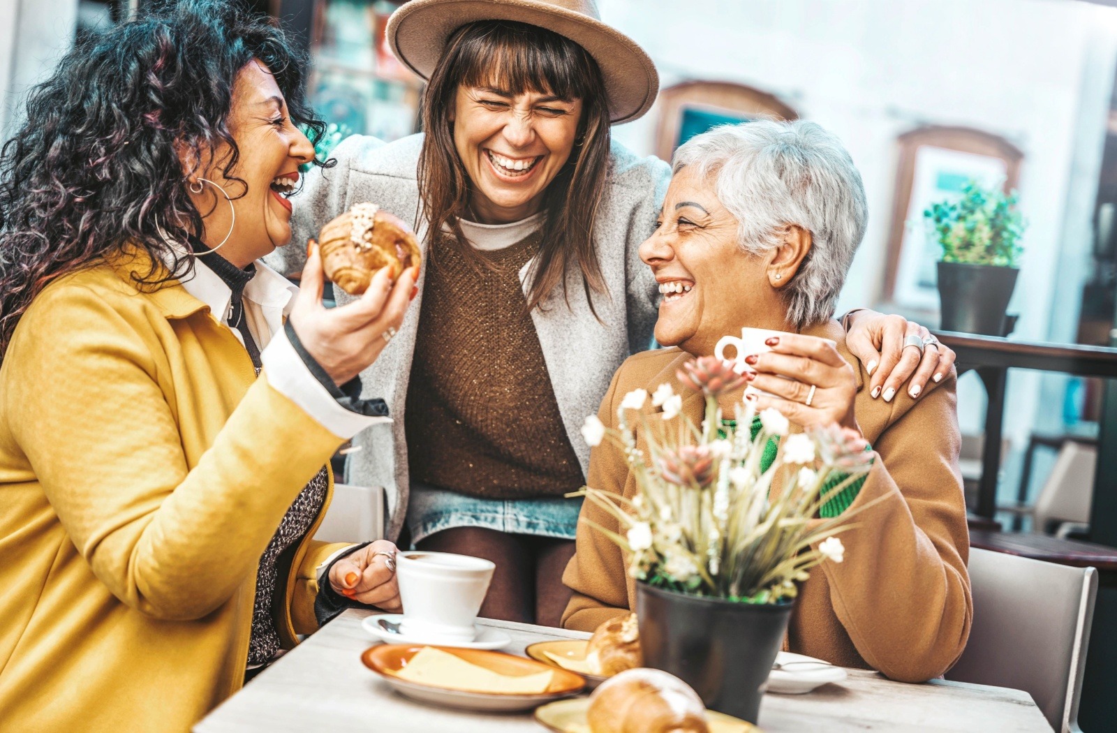 A senior, their child, and grandchild laugh together while sharing coffee and pastries.
