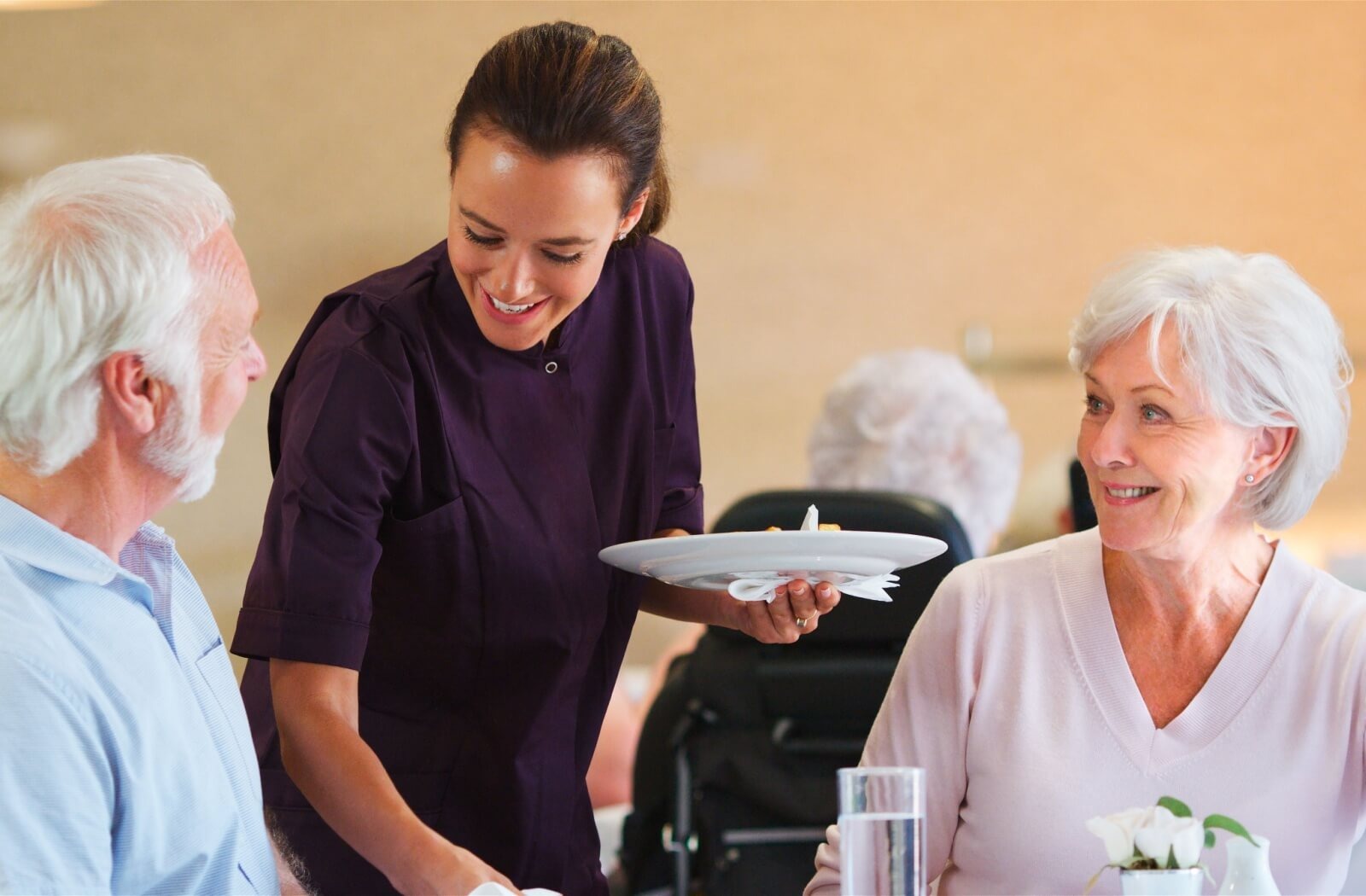 A caregiver picks up a plate from two smiling residents during mealtime in a senior living community
